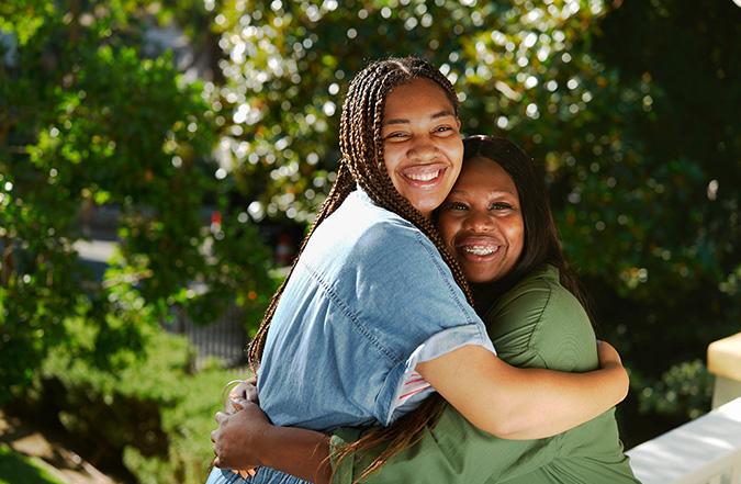 A mother and daughter smiling while they hug, with trees in the background