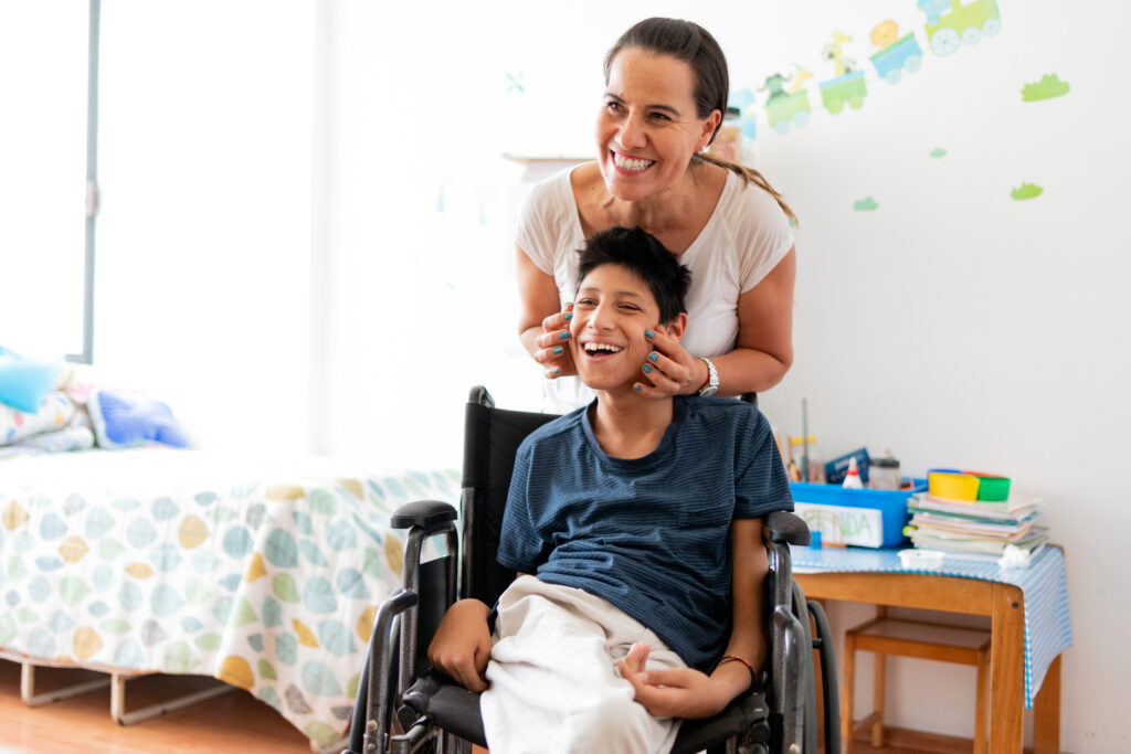 A woman stands next to and smiles with her child in a wheelchair.