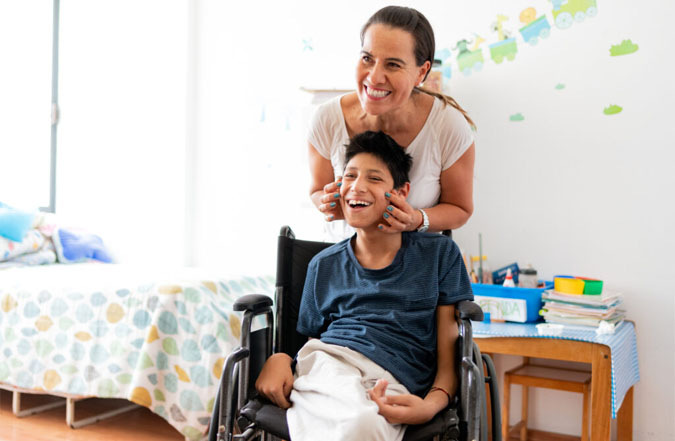 A woman stands and smiles with a smiling teenage boy in a wheelchair. 