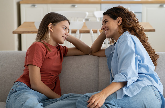 A teenaged girl and her female caregiver sit on their couch and engage in a meaningful conversation.